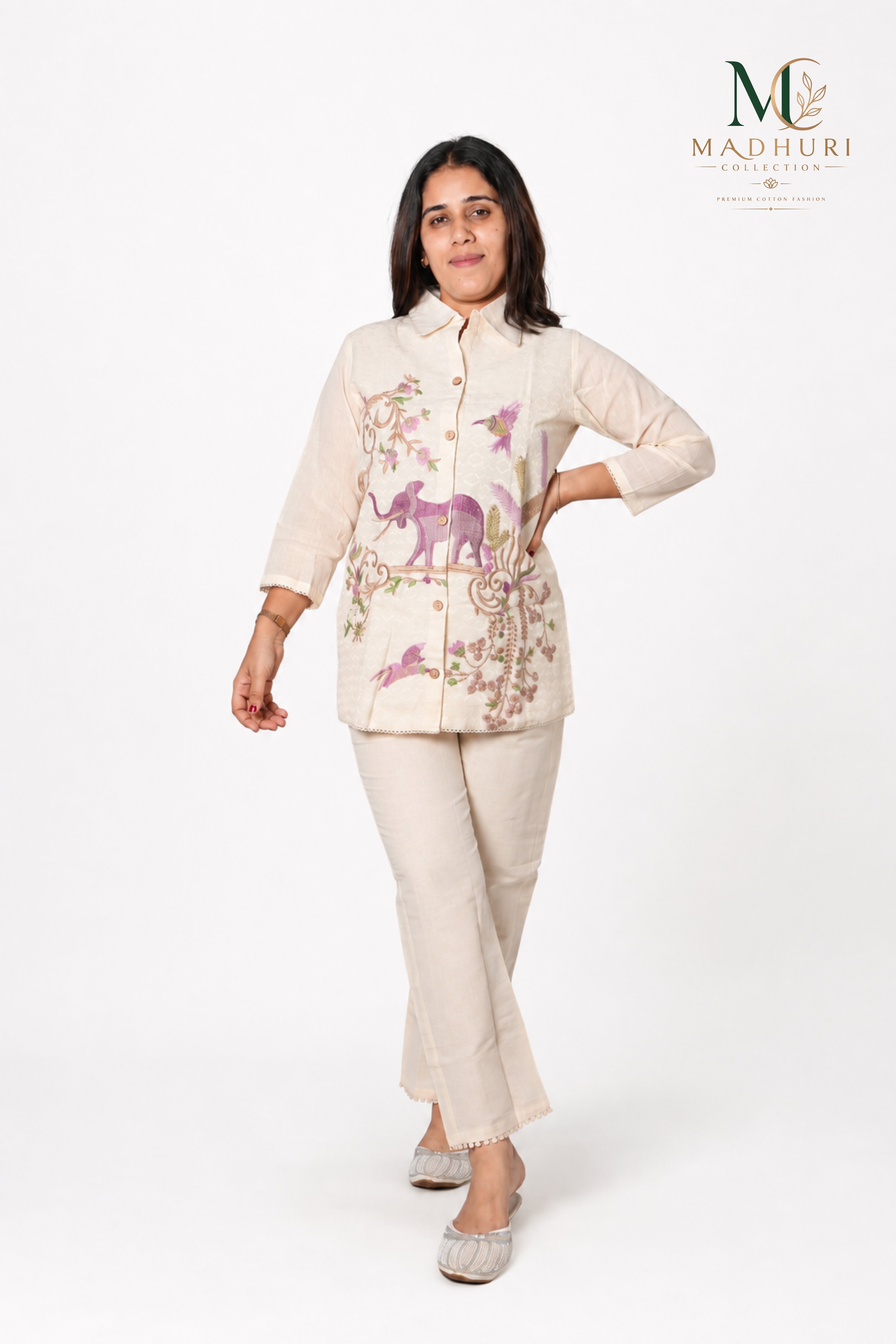 A woman modeling a cream-colored two-piece kurta set with purple and pink floral embroidery, featuring elephant and bird motifs. The set includes a long-sleeve button-up top and matching trousers, shown against a plain white studio background with the Madhuri brand logo in the top right corner.
