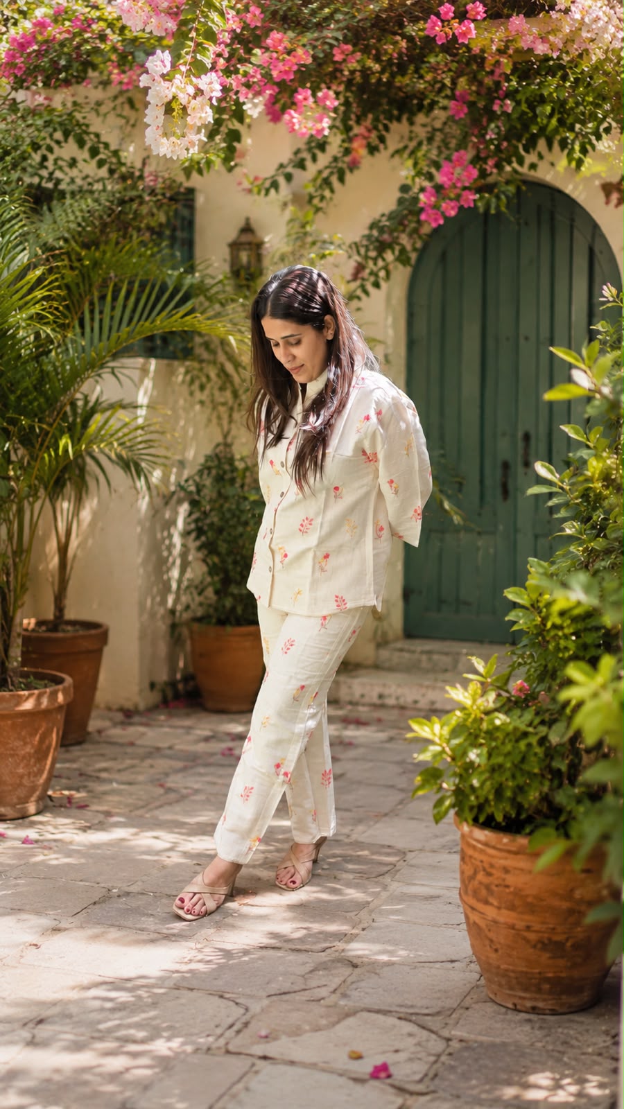 A woman wearing a cream-colored two-piece pajama set with pink floral prints stands in a garden courtyard with potted plants and a green wooden door in the background.